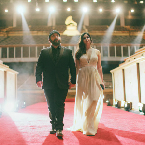 Billy Joel and Katie Lee Joel at the Grammy Awards, both dressed in formal attire. He has a beard, she has long dark hair. The venue is lit with bright stage lights and adorned with red carpet.