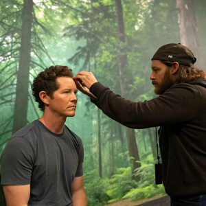 Shawn Hatosy, playing Terry McCandless, on a set break. Focused makeup artist adjusting his hair. Lush green forest backdrop