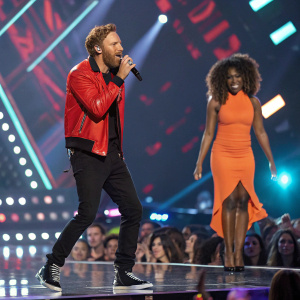 David Guetta, Caucasian male, beard, red jacket, black pants. Raye, female, African descent, curly hair, orange dress, black high heels. On stage at the MTV Music Video Awards.