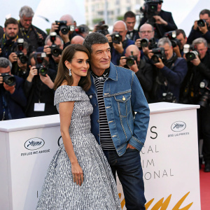 Antonio Banderas and Penelope Cruz posing for the paparazzi at Cannes Film Festival. Antonio in a stylish denim jacket, Penelope in an elegant patterned dress. Crowded red carpet scene with festival logos