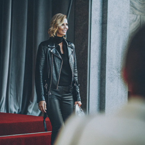 Charlize Theron at the MTV Movie Awards. Wearing a black leather jacket and black top. Dark stage backdrop with red carpet.