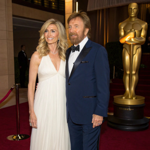 Chuck Norris and Gena O'Kelley at an award ceremony. Gena in a flowing white gown. Chuck in a navy tuxedo. Golden statue backdrop