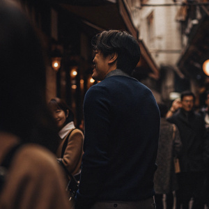 Kwon Sang-woo, wearing a casual navy sweater, stands on a vibrant city street. Crowds of people in the background.