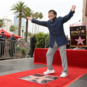 Jackie Chan celebrating the unveiling of his star on the Hollywood Walk of Fame. Wearing a navy blazer, gray trousers, and white sneakers. He poses with arms spread wide, smiling brightly. The ceremony is outdoors with palm trees and a stage in the background.