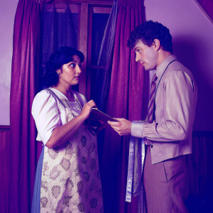 Elizabeth Peña and Daniel Hugh Kelly on set of 'I Married Dora'. Studio setting with warm lighting. Elizabeth, Hispanic woman, focused expression, standing left. Daniel, Caucasian man with curly hair, standing right. Richly colored curtain backdrop.