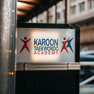 The Karoon Taekwondo Academy logo features vibrant, dynamic human figures in motion, set against a backdrop of bold red, blue, and white hues. The text is clear and prominently displayed, highlighting the academy's commitment to modern, athletic excellence.
