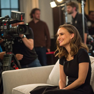 Candid moment of Natalie Portman on set. Wearing a simple black dress. Sitting on a couch with cameras around. She smiles warmly at the photographer.