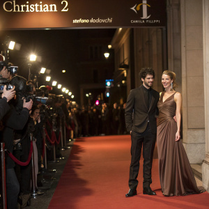 Stefano Lodovichi and Camilla Filippi at the premiere of 'Christian 2' at a luxurious cinema. Stefano in a black suit, Camilla in a brown evening gown. Red carpet event with paparazzi flashing lights
