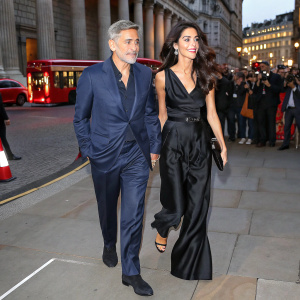 George Clooney and Amal Clooney walking out of the British Museum. Both are dressed in formal attire. George in a navy blue suit, Amal in a black satin dress. The setting is bustling London street, with people in various outfits.