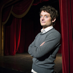 Elijah Wood, actor with curly brown hair and blue eyes, standing confidently in a dimly lit theater. Wearing a gray sweater and white shirt. The stage is set with red velvet curtains.