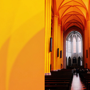 Inside the Gothic St. Michael Church. The grand hall with tall yellow-tinted pillars, high timbered ceiling, and wooden pews leading to a large altar. Soft light filters through large stained glass windows. Crowded with visitors.
