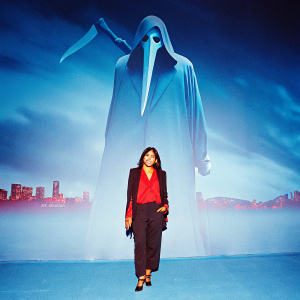 Ella-Priya D'Souza at 'Scream VI' premiere. Wearing a red blouse, black blazer, and pants. Smiling in front of a large stylized movie character with a knife-like accessory, against a backdrop of the cityscape, dark and red tones.