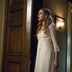 Hannah R Loyd as Diana from Grimm. She is in a white gown standing in front of a dimly lit wooden door. Scared expression