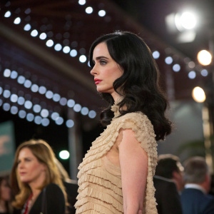 Krysten Ritter, dressed in a beige, textured, ruffled garment, with loose waves. Stands at an awards show, with ambient lighting and other celebrities around.