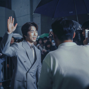 Kwon Sang-woo, in a gray suit, waving enthusiastically at a bustling convention with crowds of fans and media.