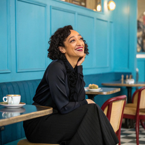 Ruth Negga, with a radiant smile, seated in a cozy cafe. Dark curly hair, black blouse and skirt. Brightly painted blue walls in the background