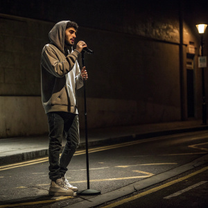 Zayn Malik, on a dimly lit street corner. Dressed casually in a hoodie. Solo performance with minimalistic backdrop.