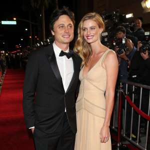Zach Braff and Taylor Bagley at a Hollywood party. Braff in a black tuxedo, Bagley in a beige dress. Red carpet with flashing cameras