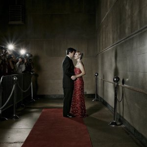 Carlo Ancelotti and Mariann Barrena McClay at a glamorous Hollywood awards ceremony. Carlo in a classic black suit, Mariann in a stunning red gown with lace details. Red carpet scene with flashing cameras and fans.