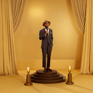 Pharrell Williams, wearing a dark suit and brown hat, giving a speech at award ceremony. Lavish stage backdrop