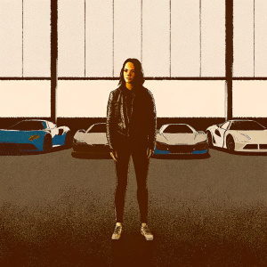 Jessica Alba, dressed in a black leather jacket, leggings, and sneakers, standing in a garage filled with racing cars. Solemn expression.