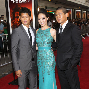 Iko Uwais, Julie Estelle, and Wayne Barrett attend the premiere of 'The Raid 2'. Movie posters and red carpet visible in background.
