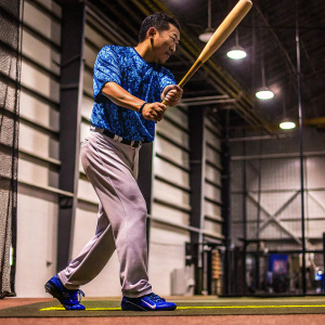 Munenori Kawasaki, Japanese baseball player, posing intensely. Gray pants, blue cleats. Blue jersey with swirling pattern. Focused stance with wooden bat. Indoor gym setting.