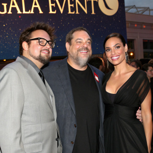 Rosario Dawson, Harvey Weinstein, and Kevin Smith attending a glamorous Hollywood gala. Kevin in a black t-shirt, full beard, and glasses. Rosario in a black gown with a plunging neckline. Harvey in a gray suit with a friendly smile. The backdrop is a shimmering, starlit sky with a banner reading 'Gala Event'.