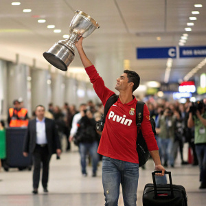 Cristiano Ronaldo celebrating at a bustling airport, hoisting the trophy aloft, dressed in casual attire with a red bwin jersey. Crowded backdrop with blurry figures.