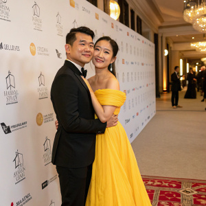 Ronny Chieng and Hannah Pham at the gala. Ronny in a black suit, Hannah in a yellow off-shoulder gown. They embrace warmly. Event hall with white backdrop and logos