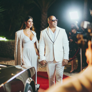 Vin Diesel and Paloma Jimenez at a Hollywood party. Both dressed elegantly, standing confidently. Bright lights and cameras in the background.