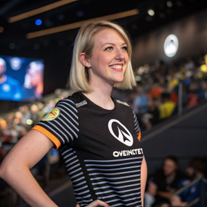 Courtney Miller, a Caucasian female with fair skin and short blonde hair, smiling confidently in a room filled with Overwatch League merchandise. She is wearing a fitted striped shirt with the Overwatch logo. The background is a blurred crowd at the event.