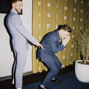 Adam Rippon and Gus Kenworthy at the Human Rights Campaign gala. Both in formal suits, posing in front of a bright red curtain with white HRC logos. Candid moment captured in a photograph
