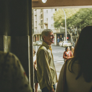 Wentworth Miller in a candid photo, standing still in a bustling cityscape. He's dressed in a casual, warm yellow shirt, wearing a small smile.