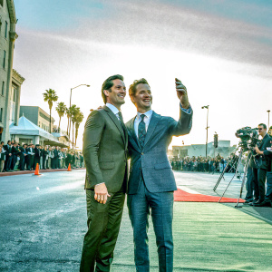 Matt Bomer and Simon Halls at the American Horror Story Hotel premiere. They're in formal wear, dark suits and ties. Friendly poses, smiling. Background is a bustling red carpet with flashing cameras and paparazzi.