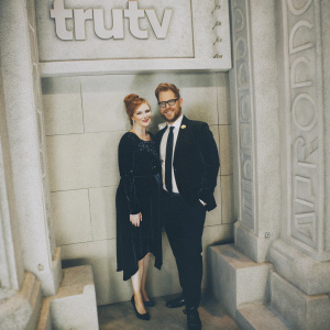 Lisa Hanawalt and Adam Conover at the TruTV after-party. Lisa in a chic black velvet dress with a bold red updo. Adam in a sharp black suit with a white shirt and dark tie. The backdrop features the TruTV logo.