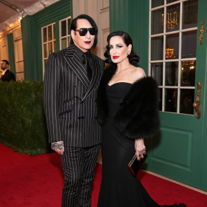 Marilyn Manson, donning a pinstripe suit, and Dita Von Teese, in a black gown with fur stole, pose elegantly at a gala event. Rich green backdrop with window details. Crimson carpet.