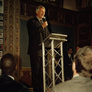 Sir Bobby Robson speaking at the Soccerex London Forum. Dressed in a black suit. Standing at a silver podium. Background features various attendees and forum branding