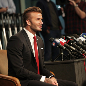 David Beckham, in a press conference. Wearing a sharp black suit and red tie. Sitting confidently.