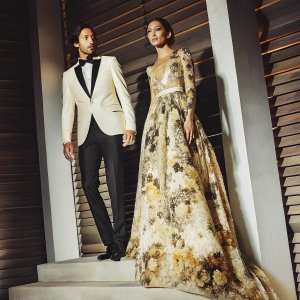 Adrien Brody and Lara Leito at the Vanity Fair Oscar Party. Brody in a white tuxedo with black bow-tie, Leito in a beige and brown floral gown. They stand on a raised platform against a stark black and white backdrop.