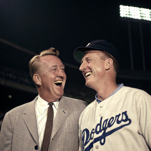 Vin Scully and Frank Howard, in vintage attire, posing for a candid shot at a Dodgers game. They are laughing together. Scully in suit and tie, Howard in Dodgers jersey. Dark stadium background.