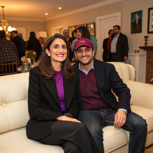 Ebru Akel and Deniz Akel in a cozy living room setting. Ebru wears a black blazer with a vibrant purple top, Deniz a dark blazer and marled cap. They are seated on a cream sofa, smiling warmly as guests mingle around them at a housewarming party.