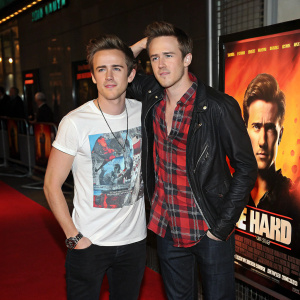 Tom Fletcher and Danny Jones from McFly attending the London Film Festival premiere of 'Die Hard 4.0'. Tom in graphic t-shirt, Danny flexing his arm in plaid shirt. They stand in front of a vibrant movie poster of the lead actor.