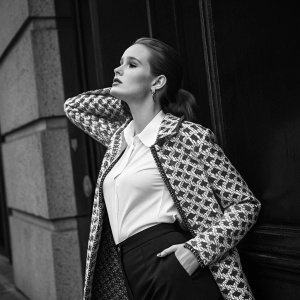 Madelaine Petsch in a black and white photoshoot. She wears a white blouse and a patterned jacket. Sophisticated pose against dark backdrop.