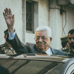 Close-up candid photo of Mahmoud Abbas, former Palestinian leader, waving to a crowd. White hair, glasses, suit and light blue shirt. Military personnel in background. High-resolution image.