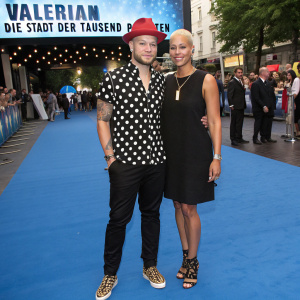 Mateo Jaschik and Tanya Ernst attending the 'Valerian - Die Stadt der Tausend Planeten' film premiere. Mateo in black and white polka dot shirt, red hat, and leopard print loafers. Tanya in sleeveless black dress and laced sandals. Stylized hairstyles. Blue carpet at CineStar.
