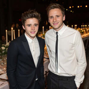 Brooklyn Beckham and Jack Ramsay at a high-profile gala. Brooklyn, with a stylish haircut and a broad smile, is in a white shirt and tie. Jack, looking casual yet elegant in a dark suit, stands beside him. A beautifully set table with candlelit centerpieces and fine dining ware lies between them.