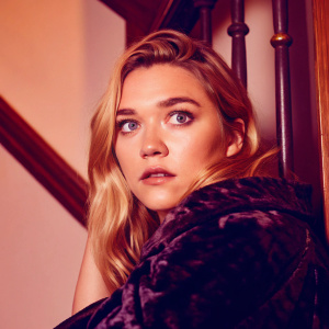 Caucasian actress Florence Pugh in a monochrome photo. Her blonde hair cascades down her shoulders. She is wearing a richly textured, dark cloak. The blurred background highlights her intense gaze.