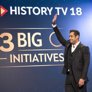 Salman Khan, in a formal suit, at the launch event of 'HISTORY TV18's 3 Big Initiatives'. Blue backdrop with the TV channel's logo and script 'HISTORY TV 18'. His hand raised in celebration.