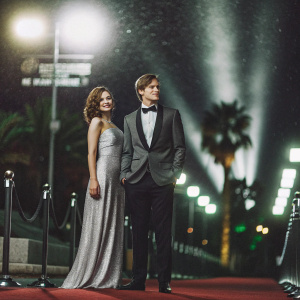 Ed Speleers and Asia Macey attending a glamorous movie festival premiere. Ed in a sharp black tuxedo, Asia in an elegant silver dress. Standing on a red carpet under a backdrop of Hollywood sign and city lights.